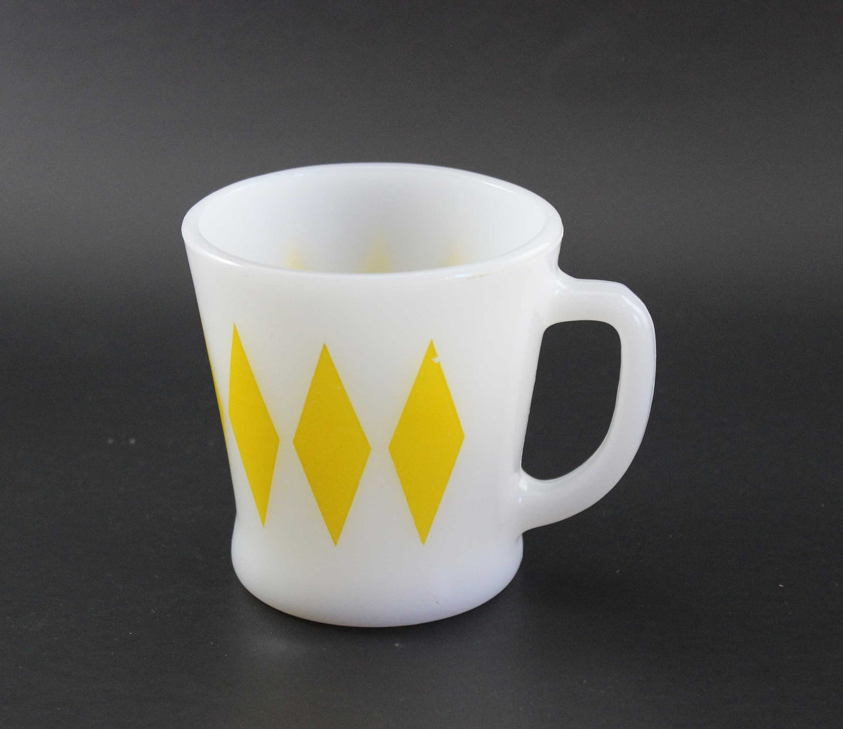 A white glass coffee mug with a yellow harlequin diamond pattern, set against a dark gray background.