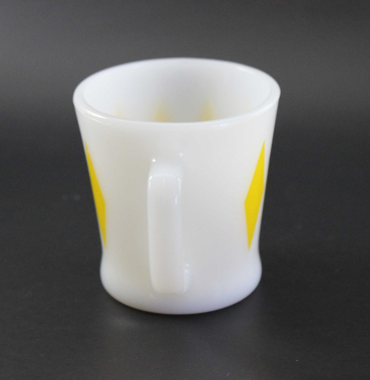 A white glass coffee mug with a yellow harlequin diamond pattern, set against a dark gray background.