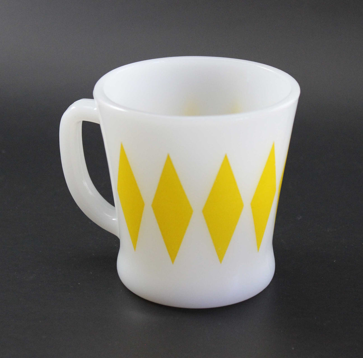 A white glass coffee mug with a yellow harlequin diamond pattern, set against a dark gray background.
