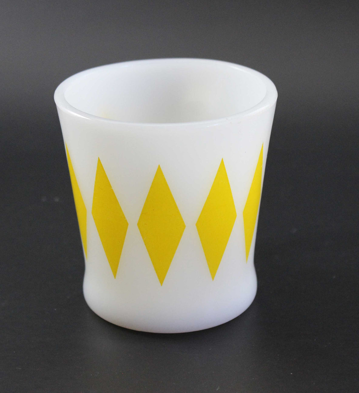 A white glass coffee mug with a yellow harlequin diamond pattern, set against a dark gray background.