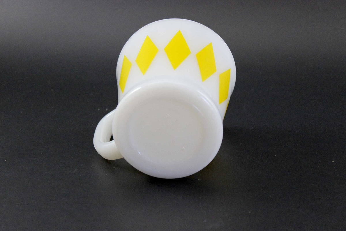 A white glass coffee mug with a yellow harlequin diamond pattern, set against a dark gray background.