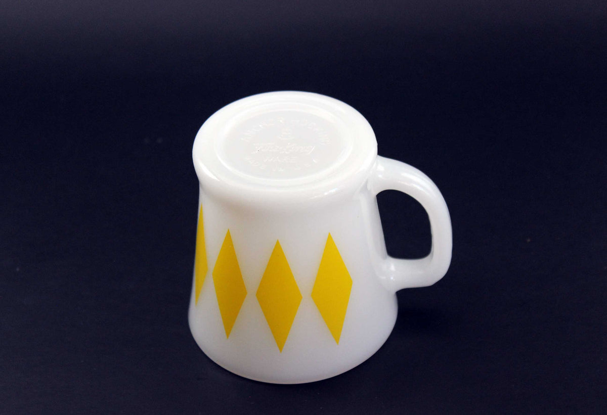A white glass coffee mug with a yellow harlequin diamond pattern, set against a dark gray background.