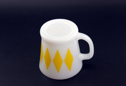 A white glass coffee mug with a yellow harlequin diamond pattern, set against a dark gray background.