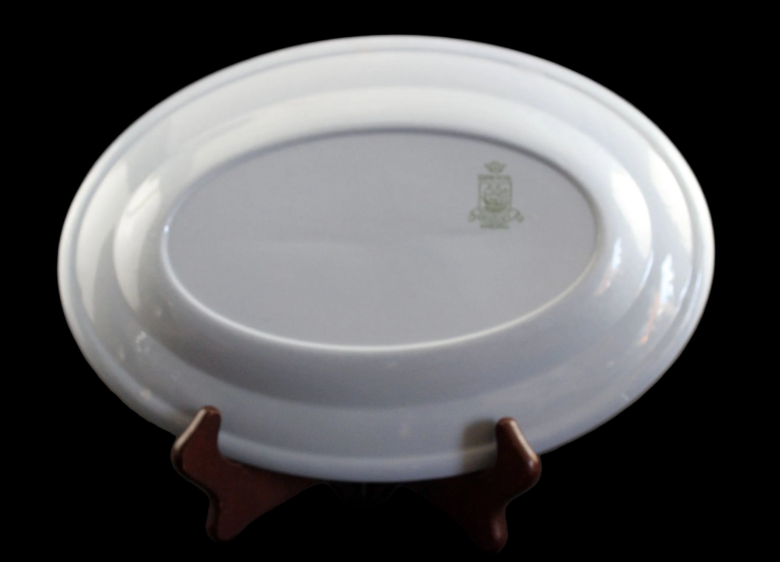An oval light blue ceramic plate displayed upside down on a small dark brown wooden stand against a dark brown background. The inside of the plate features a small light green printed crest.